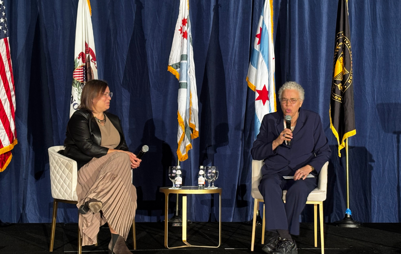 Erin Aleman and Toni Preckwinkle people sit in chairs in front of an array of flags, including the United States flag, the Illinois flag, and the Chicago flag