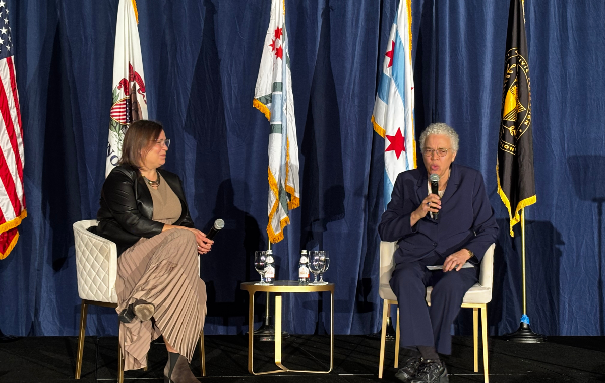 Two people sit in chairs in front of an array of flags, including the United States flag, the Illinois flag, and the Chicago flag