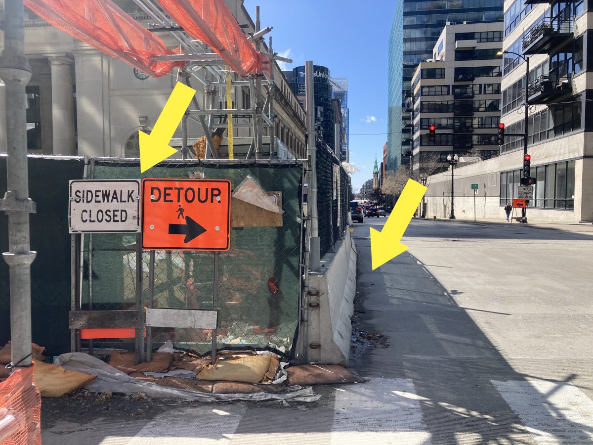 A construction site on the left with two signs in front: one sign indicates: “Sidewalk closed” and the other indicates “Detour”, with an arrow pointing to the right. To the right of the sign are concrete barriers that run between the construction and the street. One yellow arrow points to the signs as a focus indicator. A second yellow arrow points to the area next to the concrete barriers to draw focus to the area pedestrians are likely to use, which is in traffic.