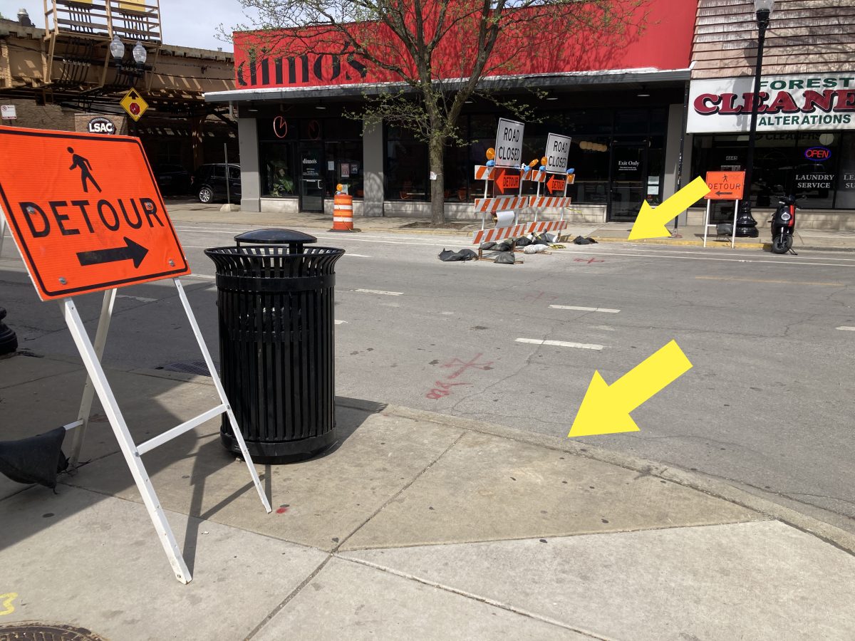 On the left is an orange construction sign placed on the sidewalk. The sign says “detour” with an arrow pointed across the street to the right. There is no curb ramp from the sidewalk on either side of the street where the detour is. Two yellow arrows point at the curbs to draw focus to the lack of curb ramps.