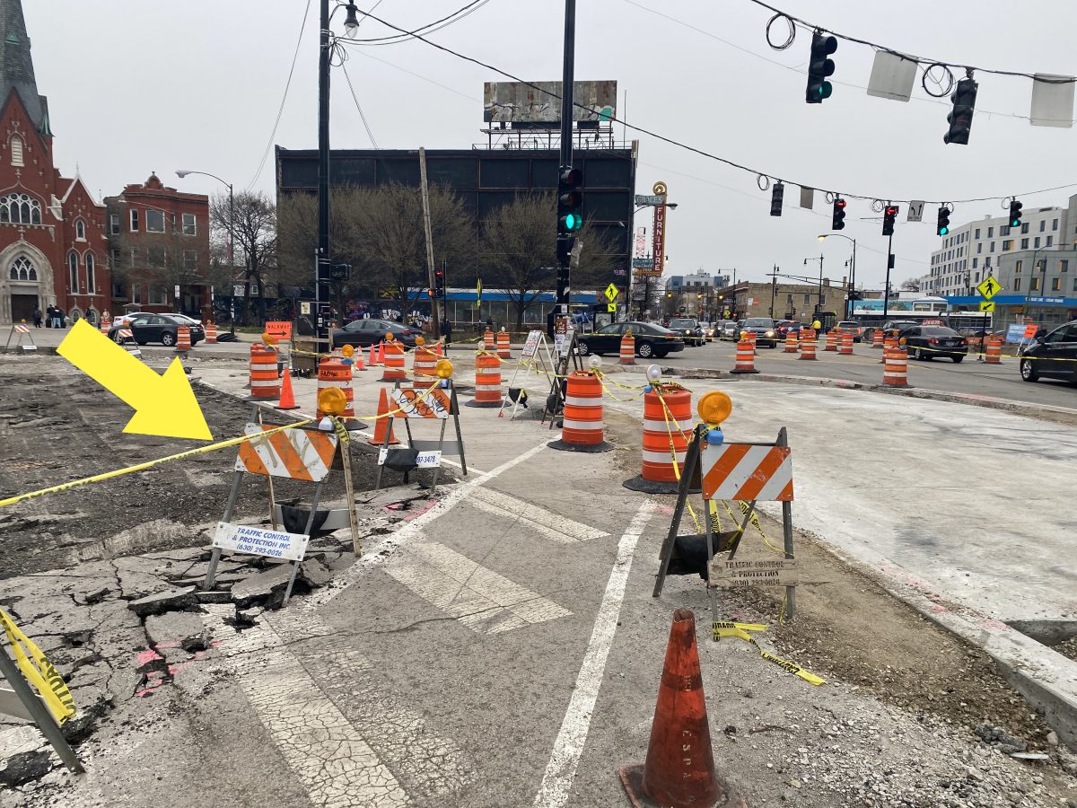 A zig-zagging asphalt path through a construction site is defined by yellow construction tape linking orange construction cones, barrels, and signs. A yellow arrow points to the yellow tape as a visual indicator. On either size of the path are rough surfaces resulting from construction