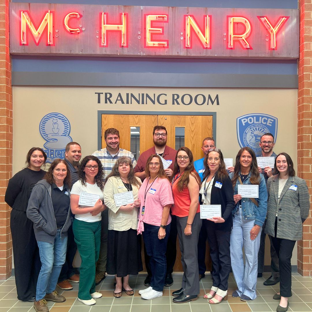 14 people, some holding certificates, stand for photo in front of training room. Neon sign saying McHenry above