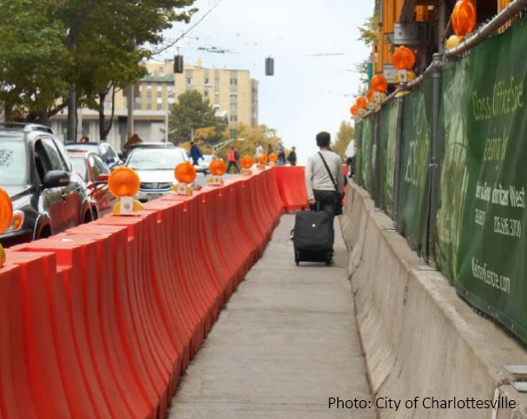 A person is walking and rolling a suitcase behind them down a temporary pedestrian path. The path defined by a hip-height orange solid barrier on the left, and a similar height concrete barrier topped with a covered chain-link fence on the right. Orange reflector lights are spaced every few feet on both barriers.