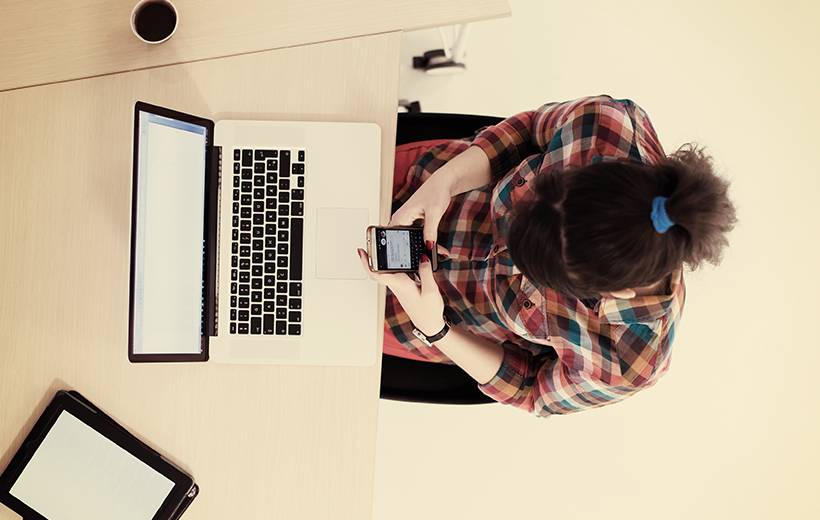 Woman sitting at desk in home office working on smartphone and laptop