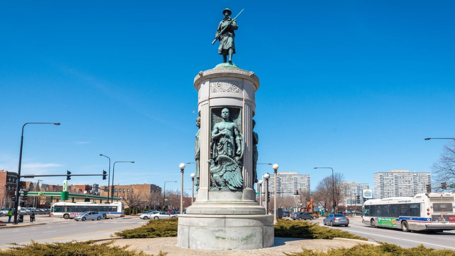 Statue on boulevard. Statue is a pillar with green copper people on three sites. Green copper figure of soldier holding gun on top of column.