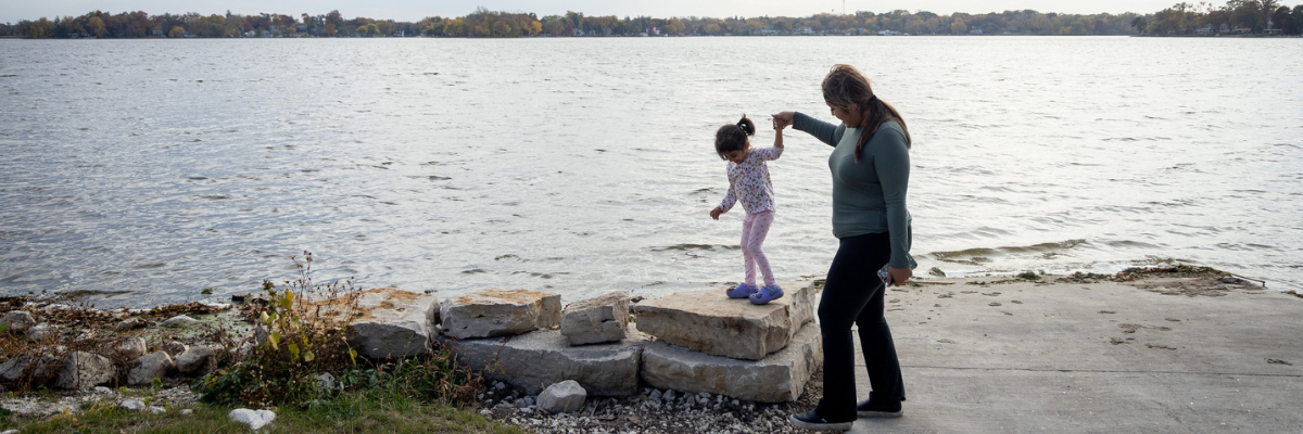 Woman holding small child's hand as she walks along rocks next to water