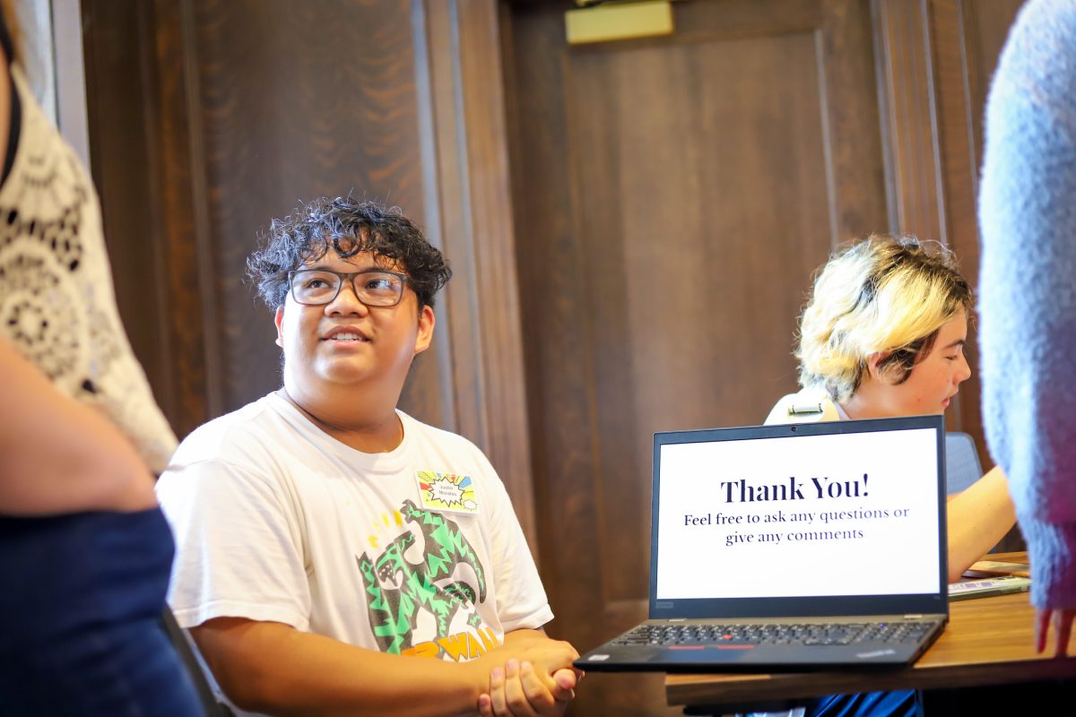 Stuend with glasses sits next to a computer and looks up at someone. The laptop screen says, "Thank you! Feel free to ask any questions or give any comments."