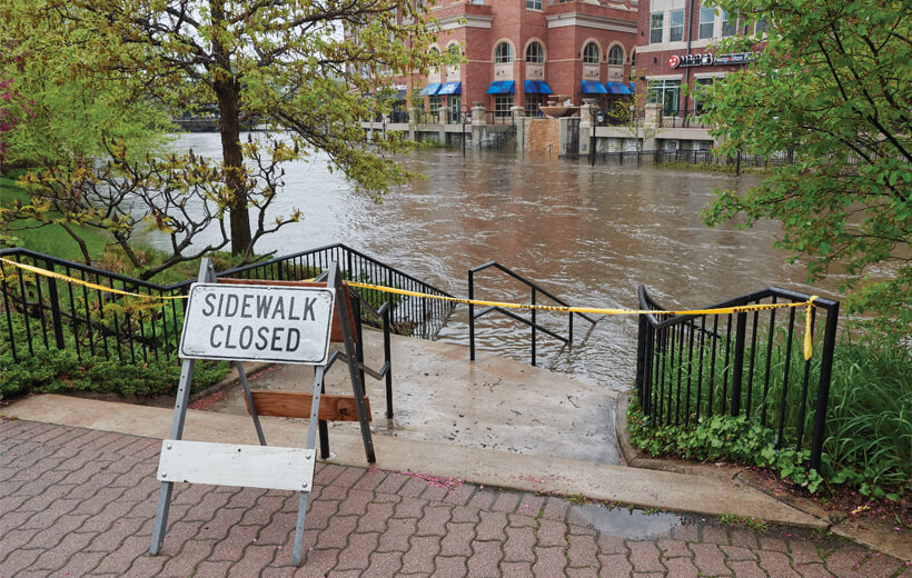 An image shows a "Sidewalk Closed" sign and yellow caution tape at the top of a staircase, with flood water submerging the bottom steps.
