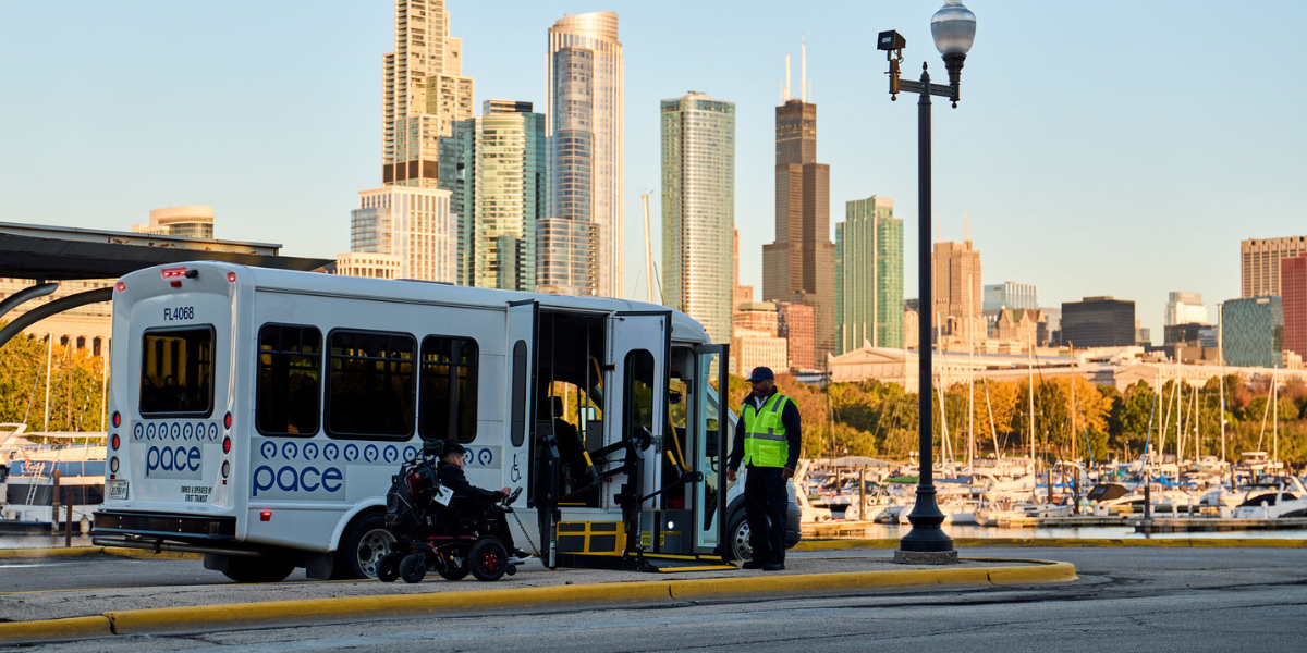A driver assists a wheelchair user boarding a Pace bus via wheelchair lift