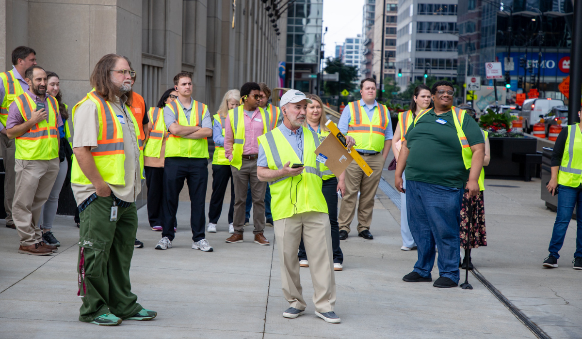 Group of people on sidewalk in high visibility vests listening to man holding microphone, clipboard, and level